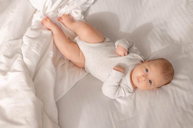 Baby with blue eyes in a white bodysuit is lying on a white bed top view