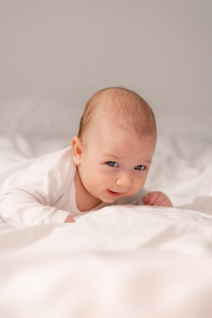 Baby with blue eyes in a white bodysuit lies on his tummy on white bed linen lifestyle