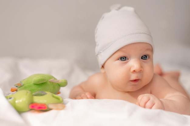 Baby with blue eyes in a white cap lying on his tummy on white bedding and playing with a green educational toy. child learns the world. lifestyle. space for text. High quality photo