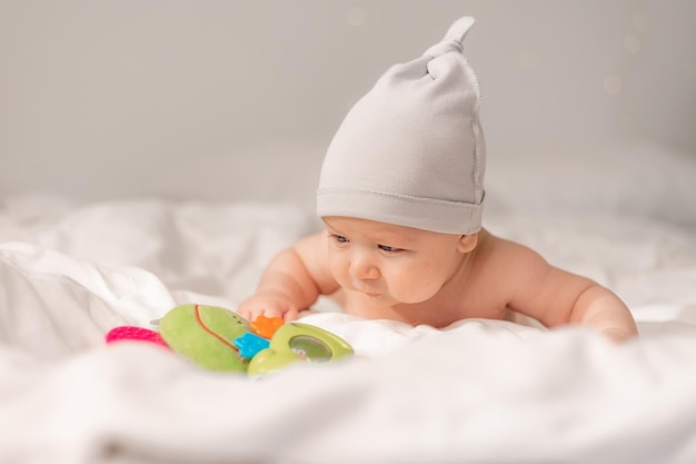 Photo baby with blue eyes in a white cap lying on his tummy on white bedding and playing with a green