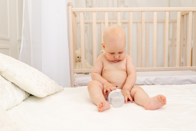 Baby with a bottle of milk sitting on the bed at home in diapers