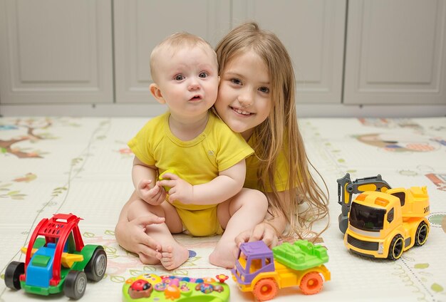 a baby in yellow clothes plays with toys and is hugged by his older sister