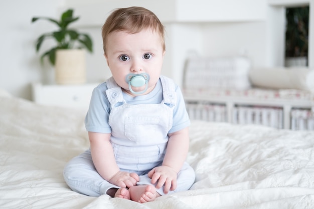 Babyjongen 6 maanden oud met tepel in blu kleren en zittend op wit bed thuis.