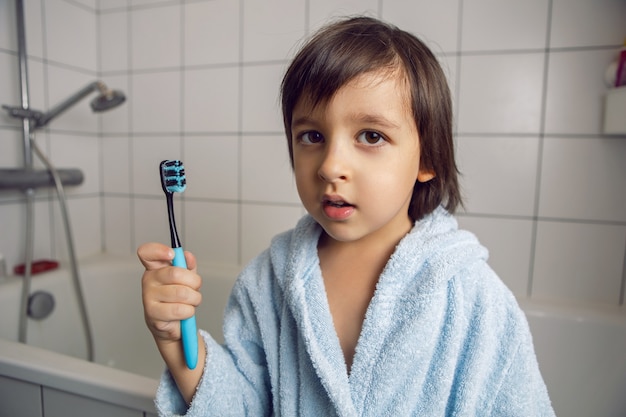 Babyjongen in een witte badkamer staat in een blauw gewaad en houdt een tandenborstel in zijn hand