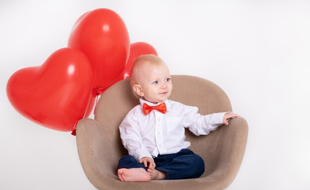 Babyjongen in pak en rode vlinderdas houdt hart ballonnen zit in stoel