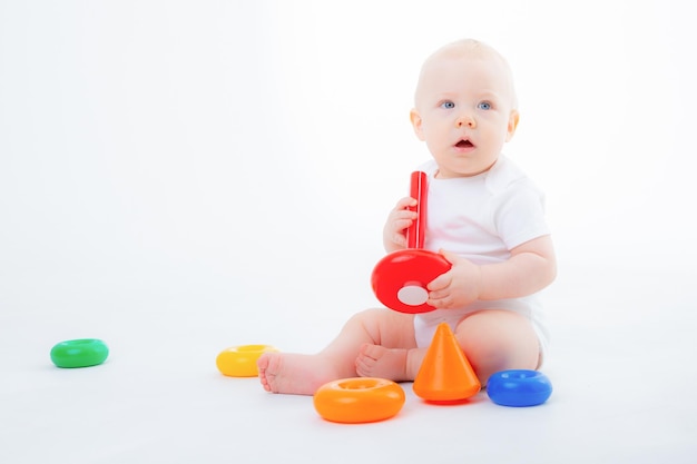 babyjongen in witte bodysuit zit te spelen met veelkleurige piramide lachend op een witte achtergrond