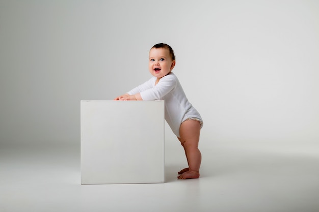 babyjongen in witte Romper staat leunend op een witte kubus op een lichte muur