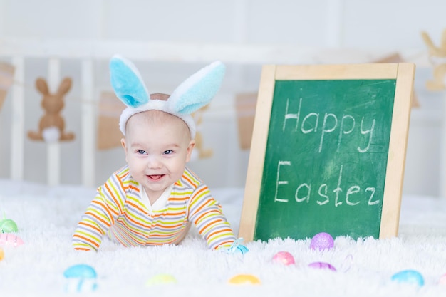 Babyjongen met konijnenoren op zijn hoofd ligt met de inscriptie Happy Easter op het bed met paaseieren schattige grappige lachende kleine baby Het concept van Pasen