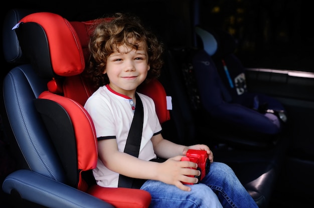 Babyjongen met krullende haarzitting in een autokinderzitje met stuk speelgoed auto in de handen