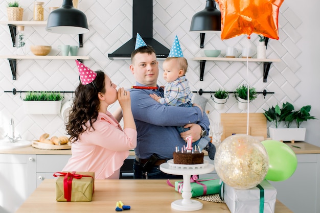 Babyjongen met ouders in heldere verjaardag hoeden viert eerste verjaardag met taart, cadeautjes, party hoorns en lucht ballonnen thuis