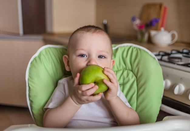 Babyjongen zit in een stoel en eet groene appel. Babyjongen eet goede voeding. Ecologische producten