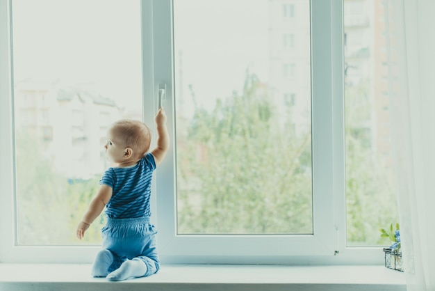 Babyjongen zit op de vensterbank en probeert het raam te openen door aan het handvat te trekken