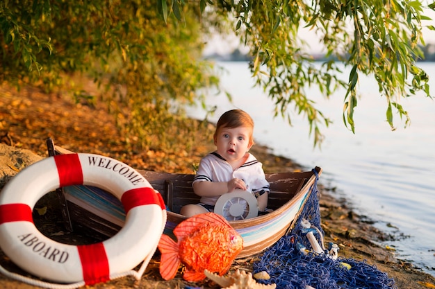 Babyjongen zittend in een boot, verkleed als zeeman, op een zandstrand met schelpen aan zee