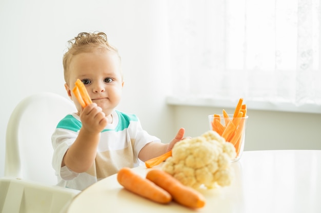 Babyjongen zittend in een kinderstoel die plakjes wortel eet op een witte achtergrond