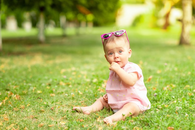 Babymeisje 7 maanden oud zittend op het groene gras in een roze Romper en heldere glazen, wandelen in de frisse lucht