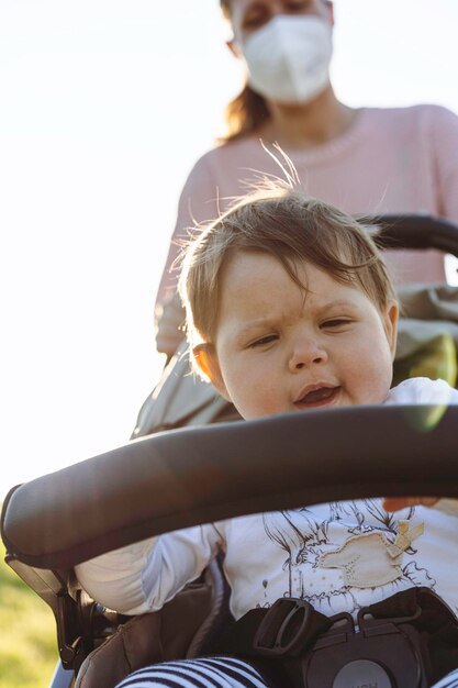 Babymeisje geniet van een wandeling in een park zittend in de moderne kinderwagen tijdens de uitbraak van het coronavirus Lief kind in buggy Moeder met beschermend gezichtsmasker