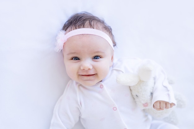 Babymeisje glimlachend op het bed op een wit katoenen bed met speelgoed dat in slaap valt of 's ochtends wakker wordt Portret van een schattige kleine baby thuis, blij en lachend