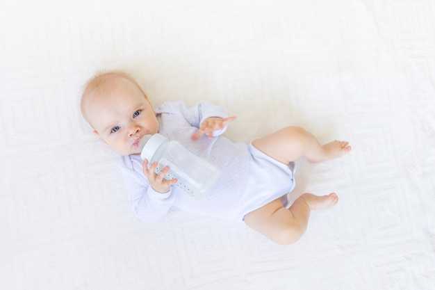 Babymeisje in een witte Romper liggend op haar rug op een wit bed thuis met een fles water