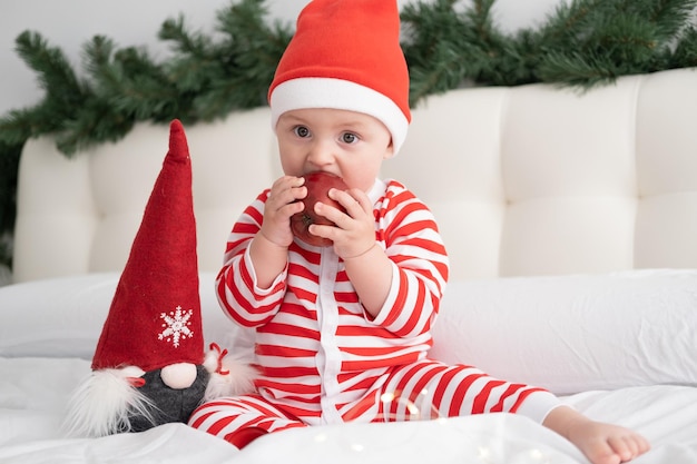 Babymeisje in gestreepte bodysuit en kerstmuts die rode appel eet op bed met kerstversiering