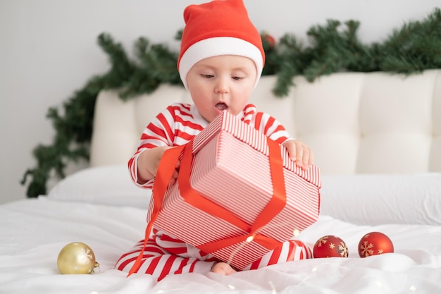 Babymeisje in gestreepte bodysuit en kerstmuts spelen met geschenkdoos op kerst versierde slaapkamer