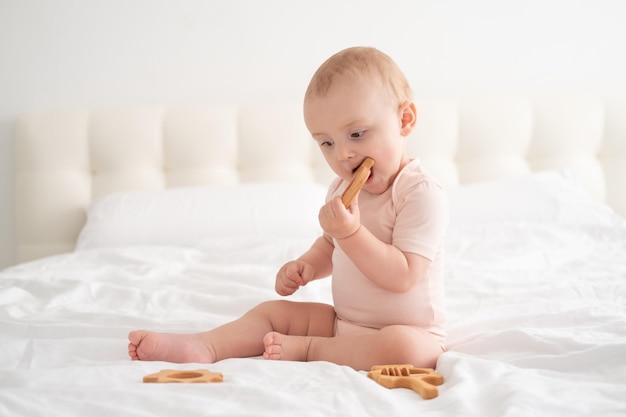 Babymeisje in lichtroze bodysuit die aan houten speelgoed knabbelt op wit beddengoed op bed