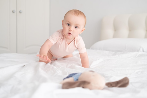 Babymeisje in lichtroze bodysuit spelen met zacht stuk speelgoed op wit beddengoed op bed