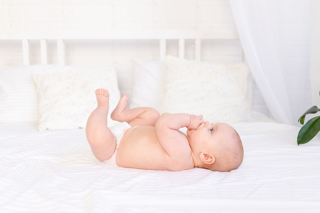 Babymeisje in luiers in een wieg op een wit katoenen bed dat op haar rug ligt en haar vuist zuigt in de kinderkamer glimlachend, pasgeboren ochtend