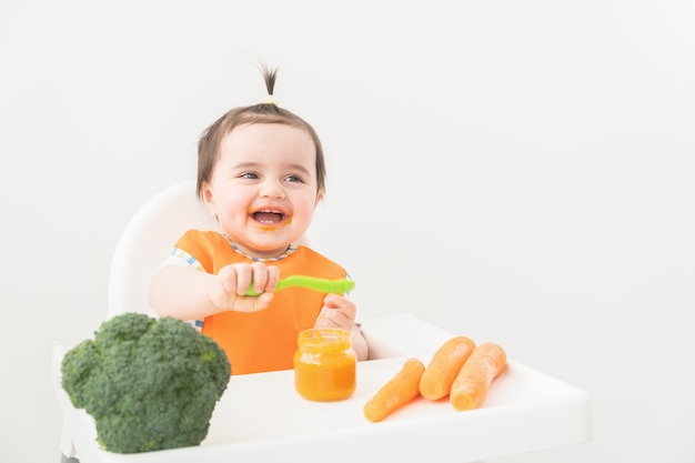 Babymeisje in oranje slabbetje zittend in een stoel Childs plantaardige puree eten.