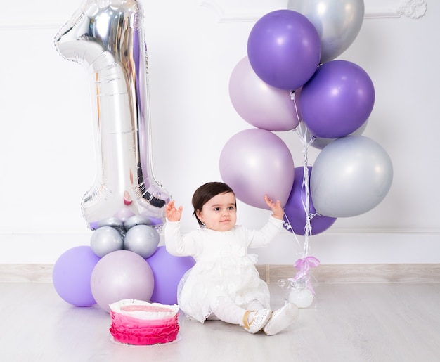 Babymeisje in witte jurk zittend op de vloer viert haar eerste verjaardag met cake en ballonnen.