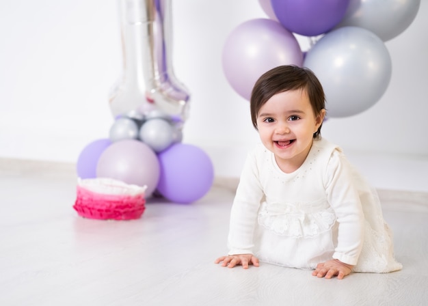 Babymeisje in witte jurk zittend op de vloer viert haar eerste verjaardag met cake en ballonnen.