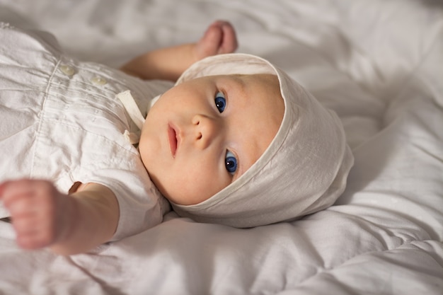 Babymeisje met blauwe ogen in witte jurk en pet in bed.