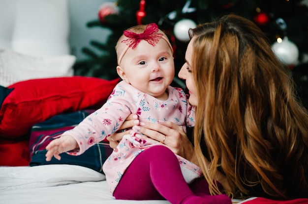 Babymeisje met moeder op bed in de slaapkamer in de buurt van de kerstboom. Vrolijk kerstfeest. Kerst versierd interieur. Het concept van gezinsvakantie.