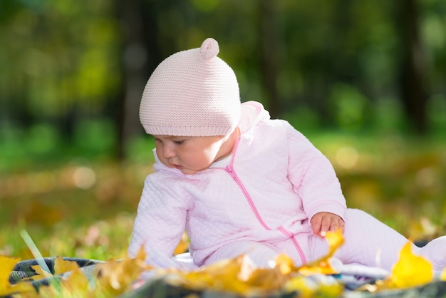 Babymeisje spelen met herfstbladeren als ze zit op een deken op het gras in een park in een roze springpak en hoed