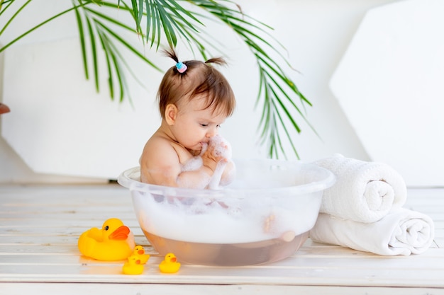 Babymeisje wast en speelt in een bak met schuim en water in een lichte kamer thuis