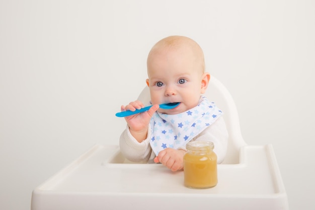 Babymeisje zit op een kinderstoel, houdt een lepel vast en eet fruitpuree