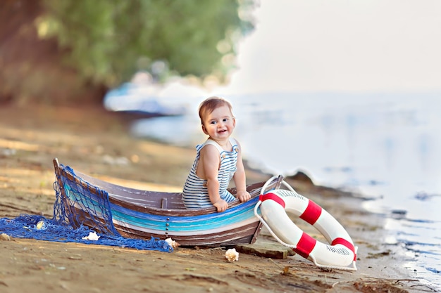 Babymeisje zittend in een boot, verkleed als zeeman, op een zandstrand met schelpen aan zee