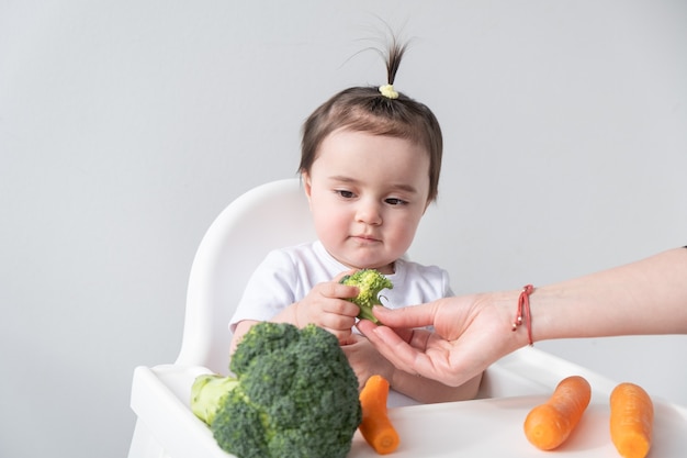 Babymeisje zittend in een kinderstoel wortel en broccoli eten.