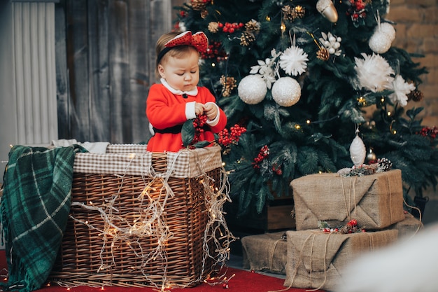 Babyvrouw die op vloer met Kerstmisbal kruipen die de hoed en het kostuum van de Kerstman over lichten dragen bij achtergrond