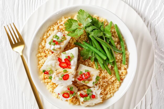 Bacalhau com todos cod with bulgur steamed green beans sprinkled with hot chili peppers and spring onions in a white bowl on a wooden table balkan cuisine flat lay