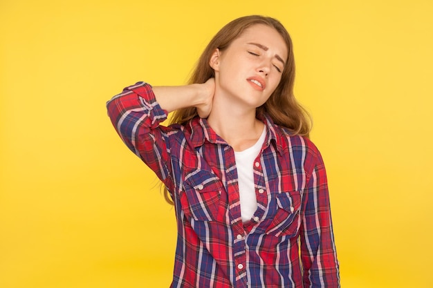 Back ache. Portrait of sick unhealthy girl in checkered shirt touching sore neck, grimacing from pain, massaging spine, feeling fatigued exhausted. indoor studio shot isolated on yellow background