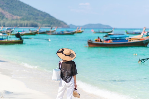 Back of asian woman wearing shirt and a hat with holding shoe\
on a sunny beach with sea and boats in the background concept of\
holidays and travel picture looking alone or sad she looking\
forward