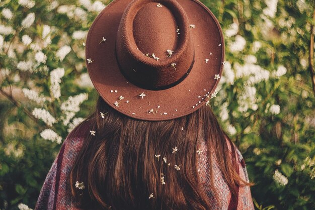 Photo back of beautiful hipster girl in hat and poncho with amazing hair with flowers standing in park in evening sunshine stylish woman traveler in spring garden space for text atmospheric moment