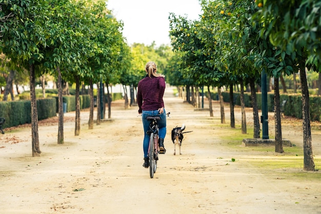 Back of a blonde woman riding a bicycle while walking the dog in a public park