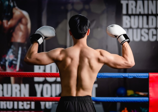 Задняя часть тела молодого человека без рубашки в белых боксерских перчатках, поза стоя, поднимает руки вверх, чтобы показать мышцы спины на боксерском ринге в фитнес-зале,