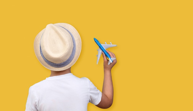 The back of a boy in a hat holding a plane in his hands on a yellow background. Copy space. Summer vacation concept.