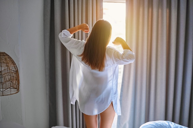 Back of brunette young girl stretching after waking up in the morning and standing at sunbeam window