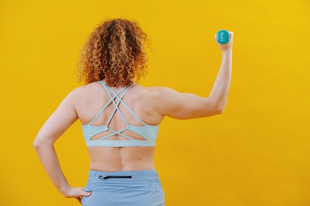 Back image of a young woman pumping her biceps with a tiny\
dumbbell