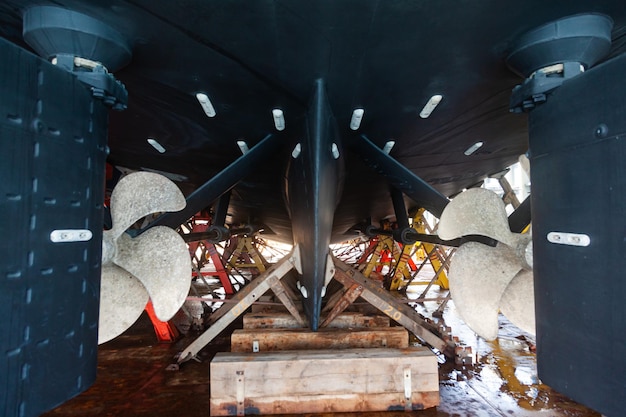 The back of a large motor yacht with two propellers standing on wooden blocks in dry dock
