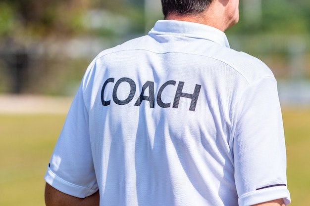 Back of male sport coach wearing COACH shirt at an outdoor sport field