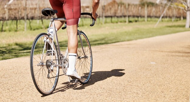 男性の自転車と公園のパスの後ろ、日差しの中でのトライアスロン スポーツの運動とトレーニング、自由なカーディオ フィットネス アドベンチャーとパフォーマンスのモックアップのためのサイクリストの脚の自転車と屋外サイクリング トレイル